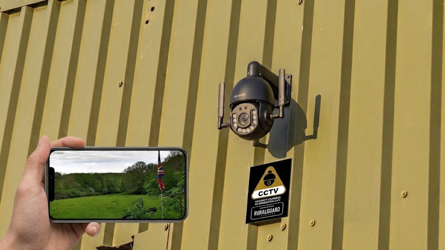 A PTZ CCTV security camera mounted on a yellow corrugated metal wall, with a hand holding a smartphone in the foreground showing the camera's live feed of a green landscape.