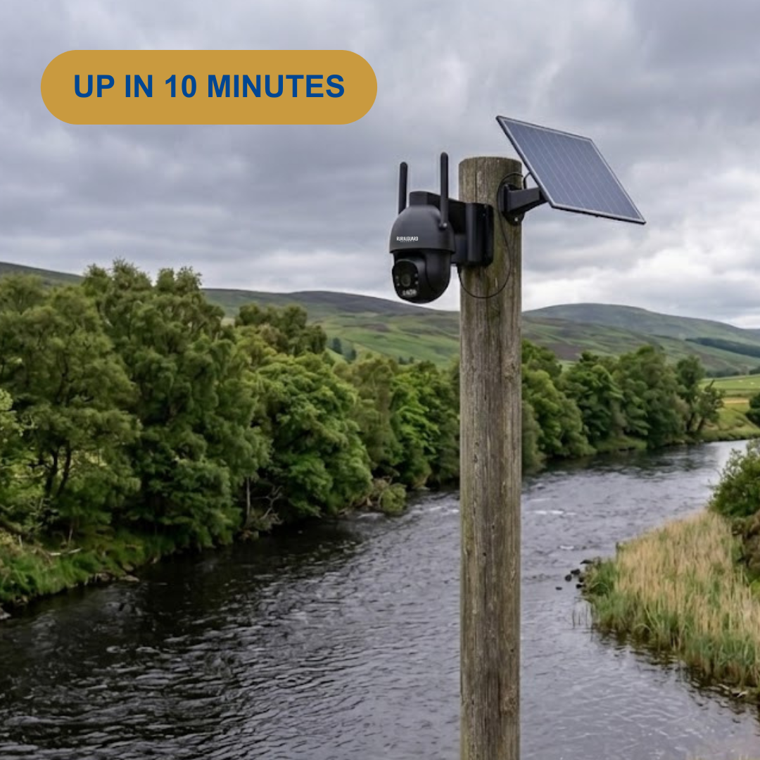 A black wireless security camera mounted on a wooden post with an attached solar panel, set outdoors beside a river with trees and hills in the background. An orange badge at the top left reads 'UP IN 10 MINUTES'.